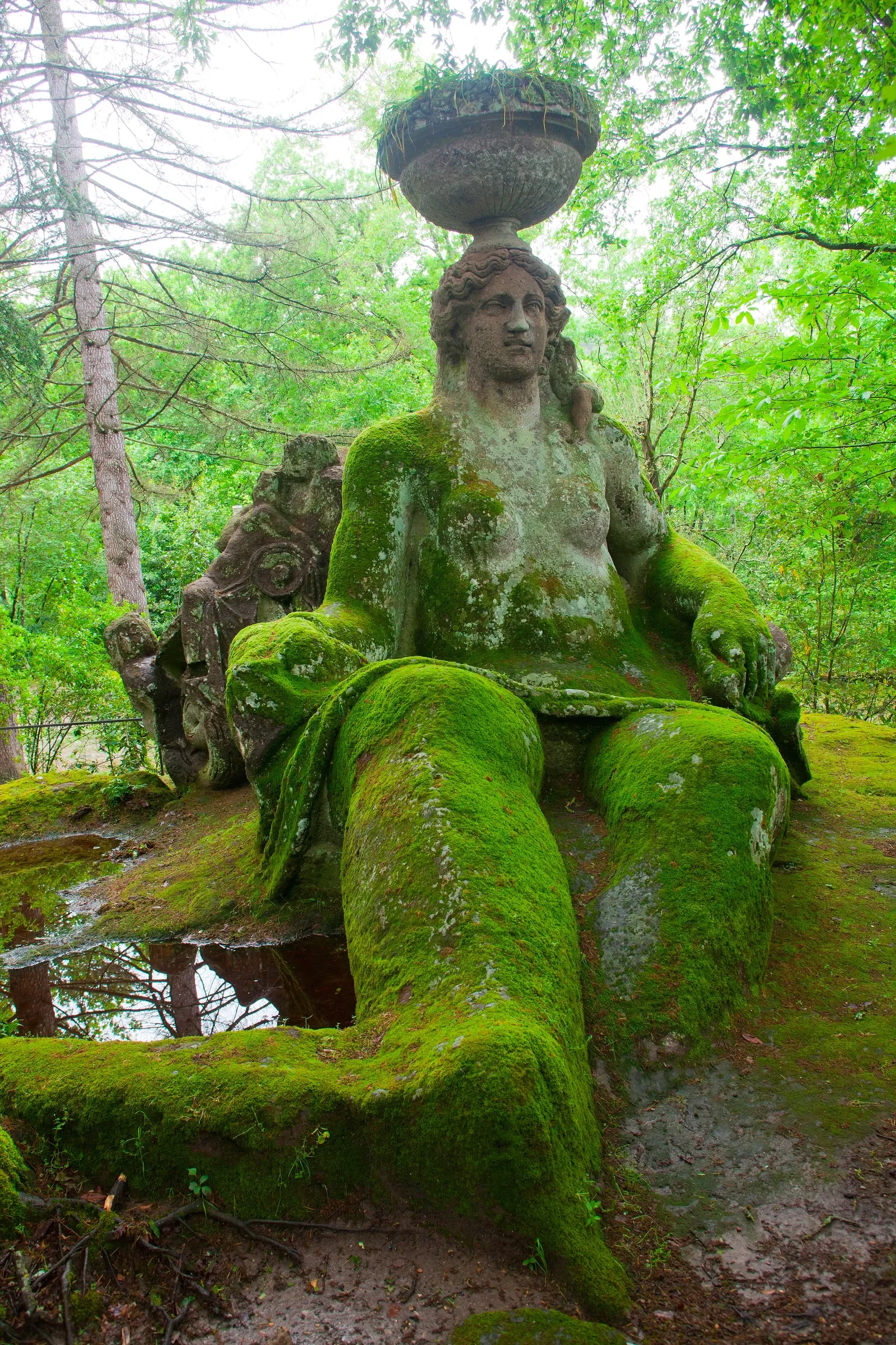 Statua di una figura femminile seduta con un cappello sopra la testa, ricoperta di muschio, in una foresta verde