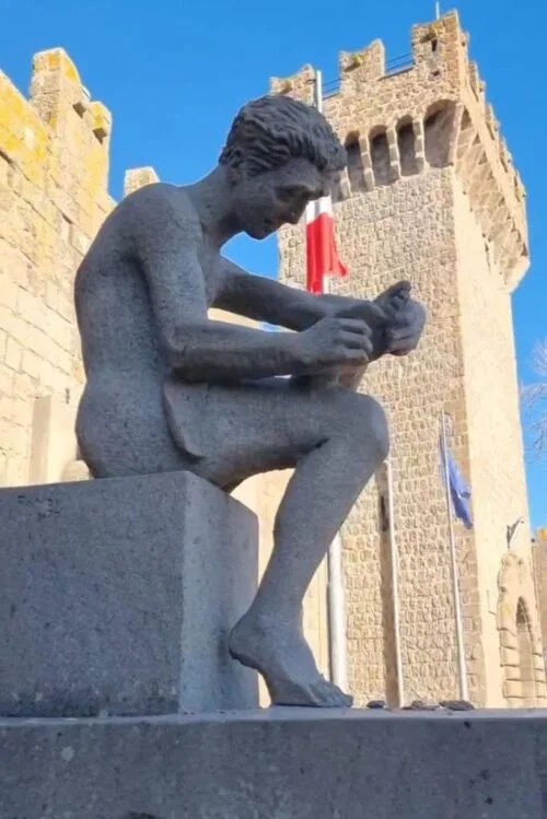 Statua di un ragazzo seduto con un libro in mano, in fronte a un castello medievale con bandiere.