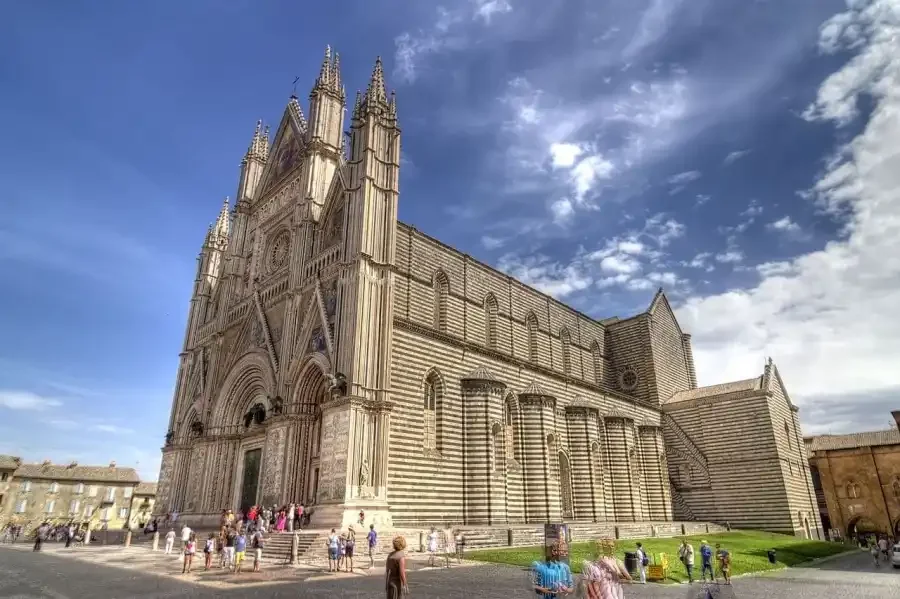 Cattedrale gotica in pietra con facciata decorata e torri alte, con persone davanti, cielo nuvoloso.
