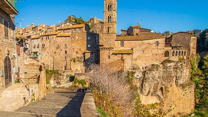Paesaggio di un insediamento storico con edifici in pietra, una torre, e una scalinata, su una scogliera rocciosa