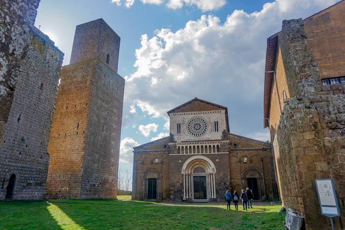 Chiesa medievale con facciata in pietra e tre persone davanti, circondata da antiche mura.