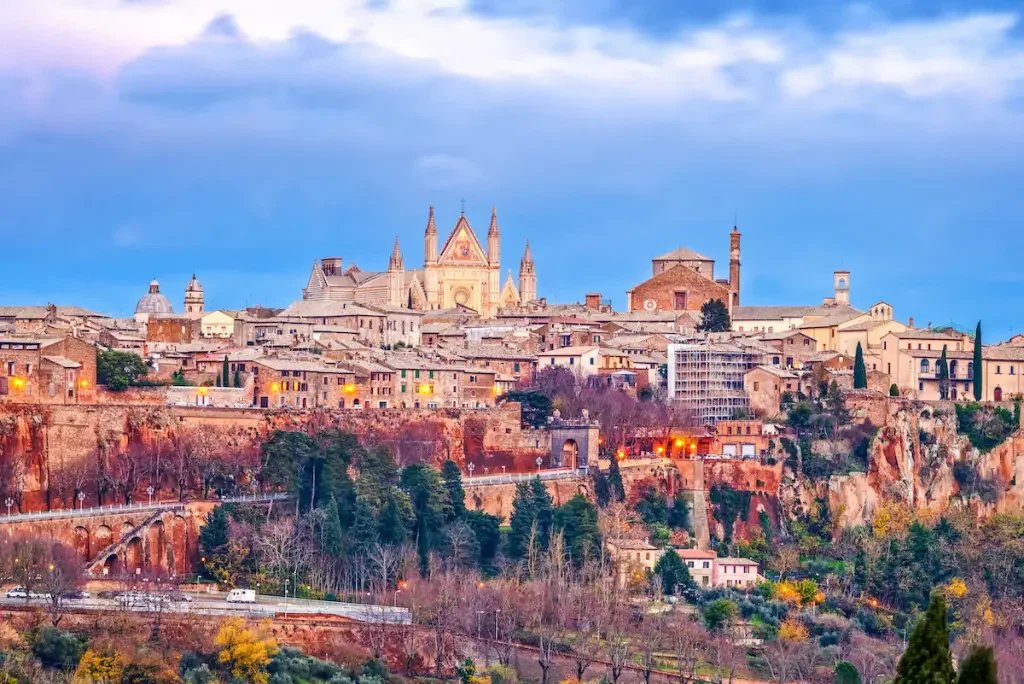 Vista di una città medievale con un grande edificio religioso in cima, su una collina, al tramonto.