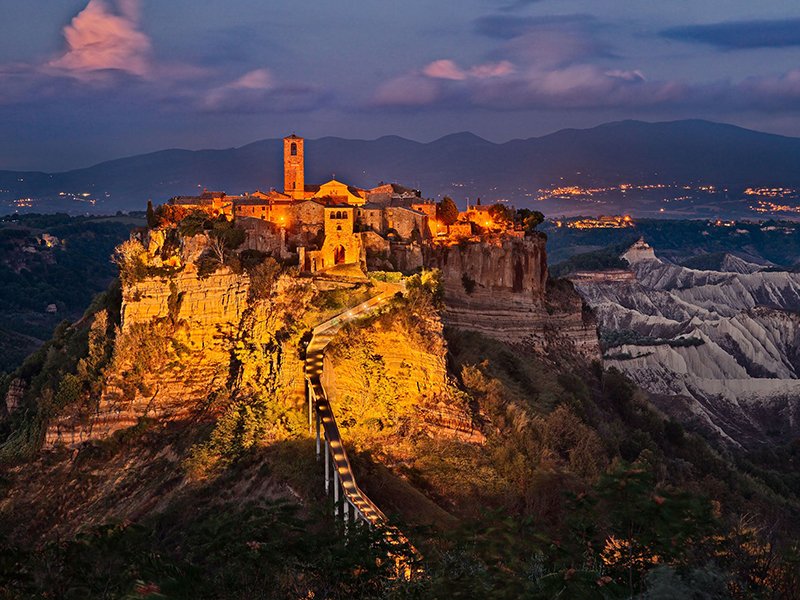 Paesaggio notturno con un borgo storico su una collina illuminato, con montagne sullo sfondo e un cielo crepuscolare.