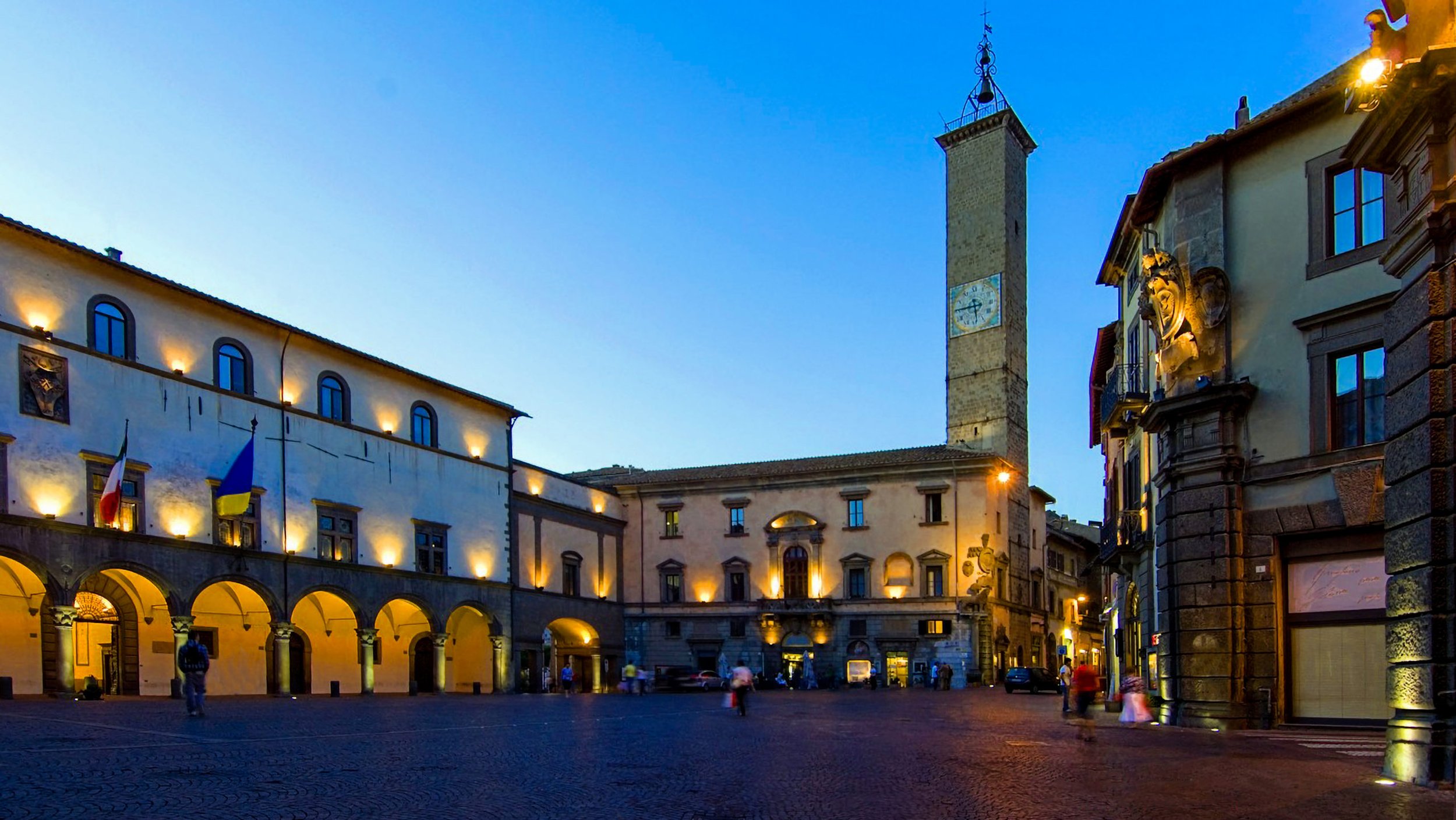 Una piazza con edifici storici illuminati durante la sera, con una torre con orologio e alcune persone che camminano.