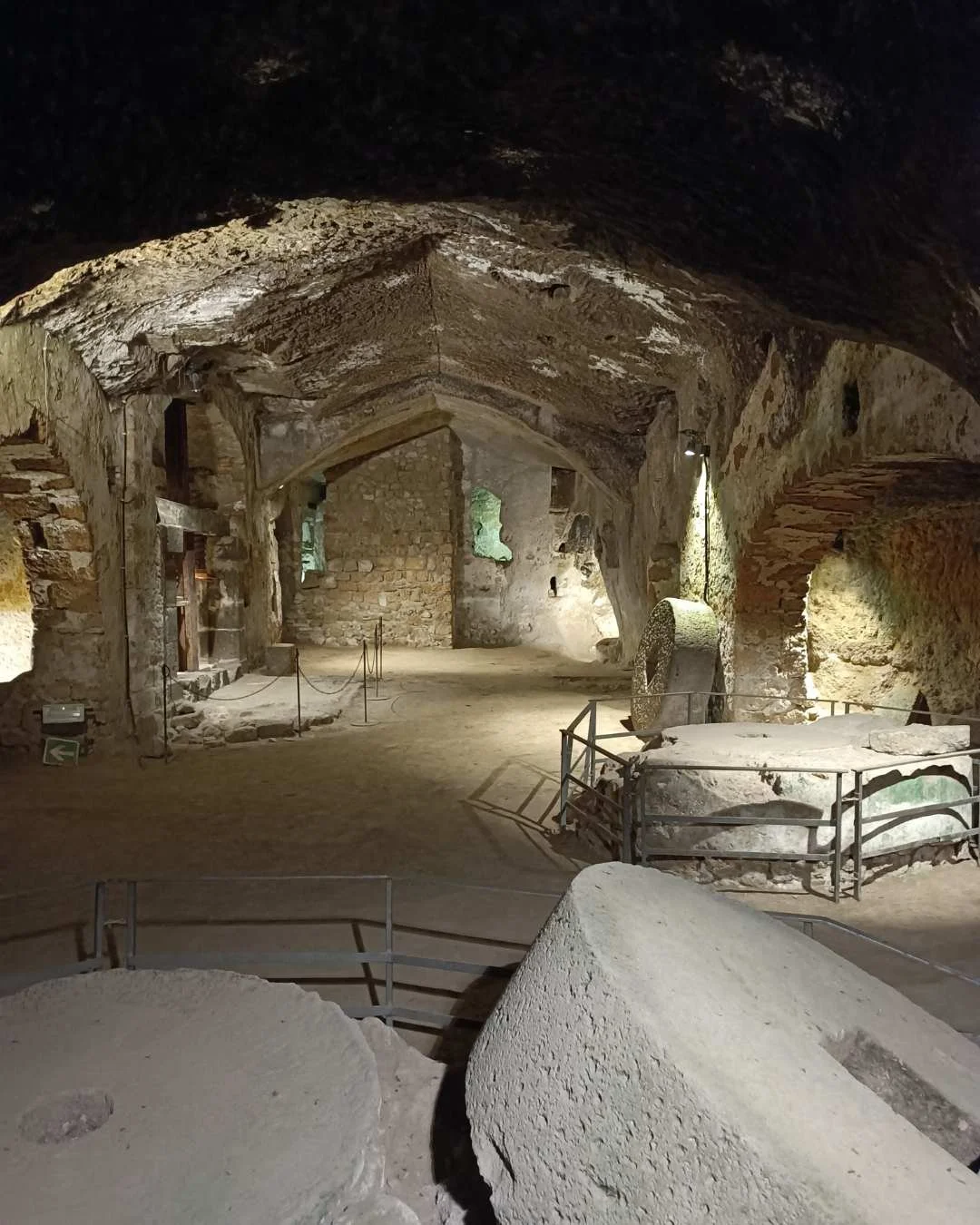 Interno di una grotta con pareti di pietra, alcune macine di pietra e strutture di supporto in metallo, illuminate da luci artificiali.