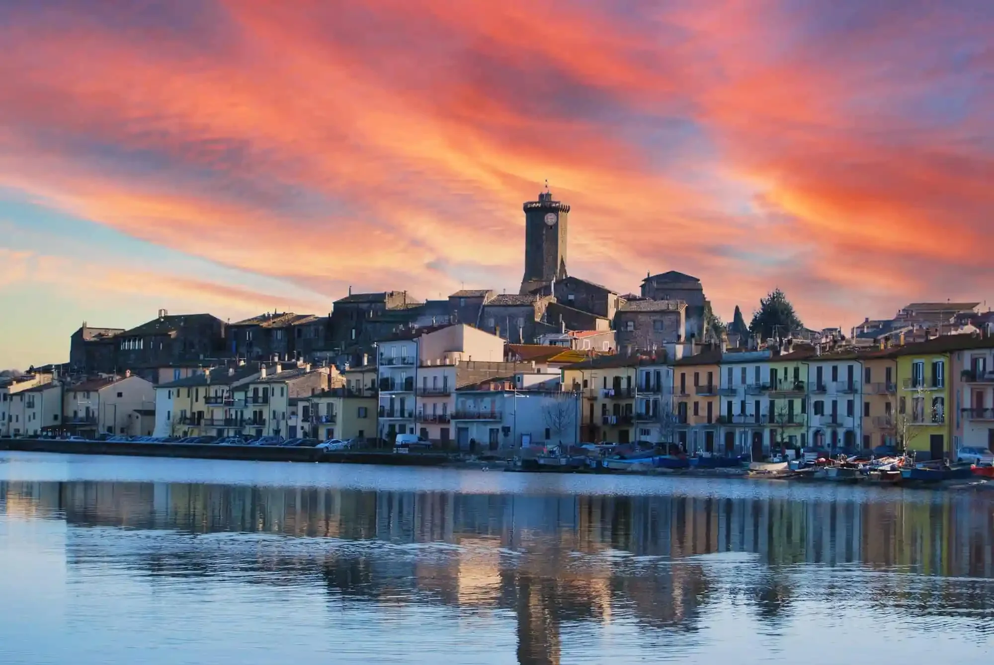 Un paesino di case colorate sul lungolago, con un castello o torre con orologio sulla collina sullo sfondo, sotto un cielo al tramonto con nuvole rosa e arancioni.