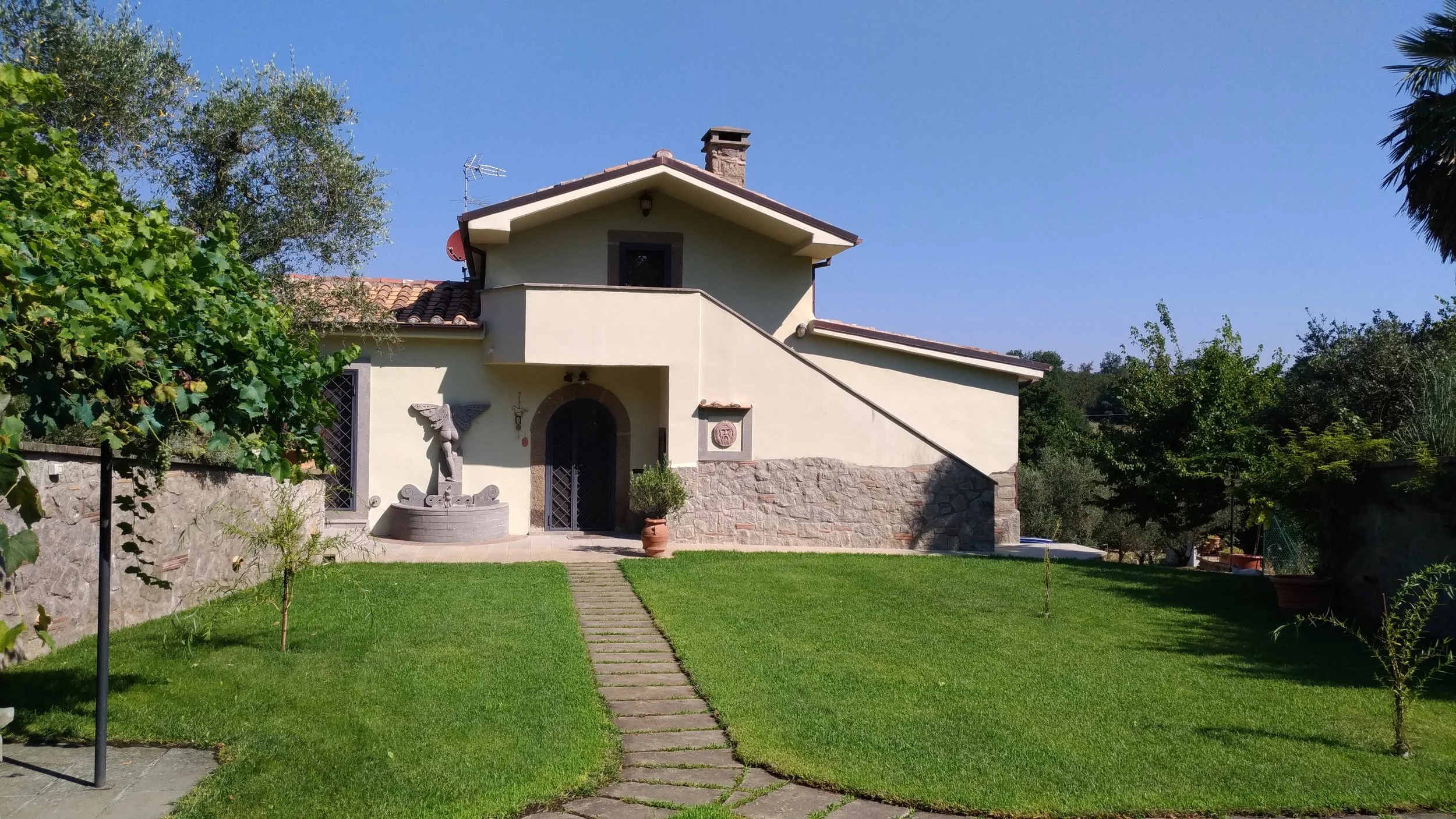 Casa di campagna con giardino ben curato, al centro un sentiero di pietra, al lato un busto decorativo e piante verdi, sotto un cielo blu chiaro.
