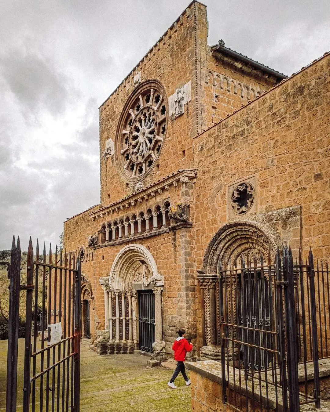 Facciata di una chiesa storica in pietra con grandi portali gotici, una finestra rotonda con vetrate e un grande rosone. Un bambino con giacca rossa cammina davanti alla chiesa.