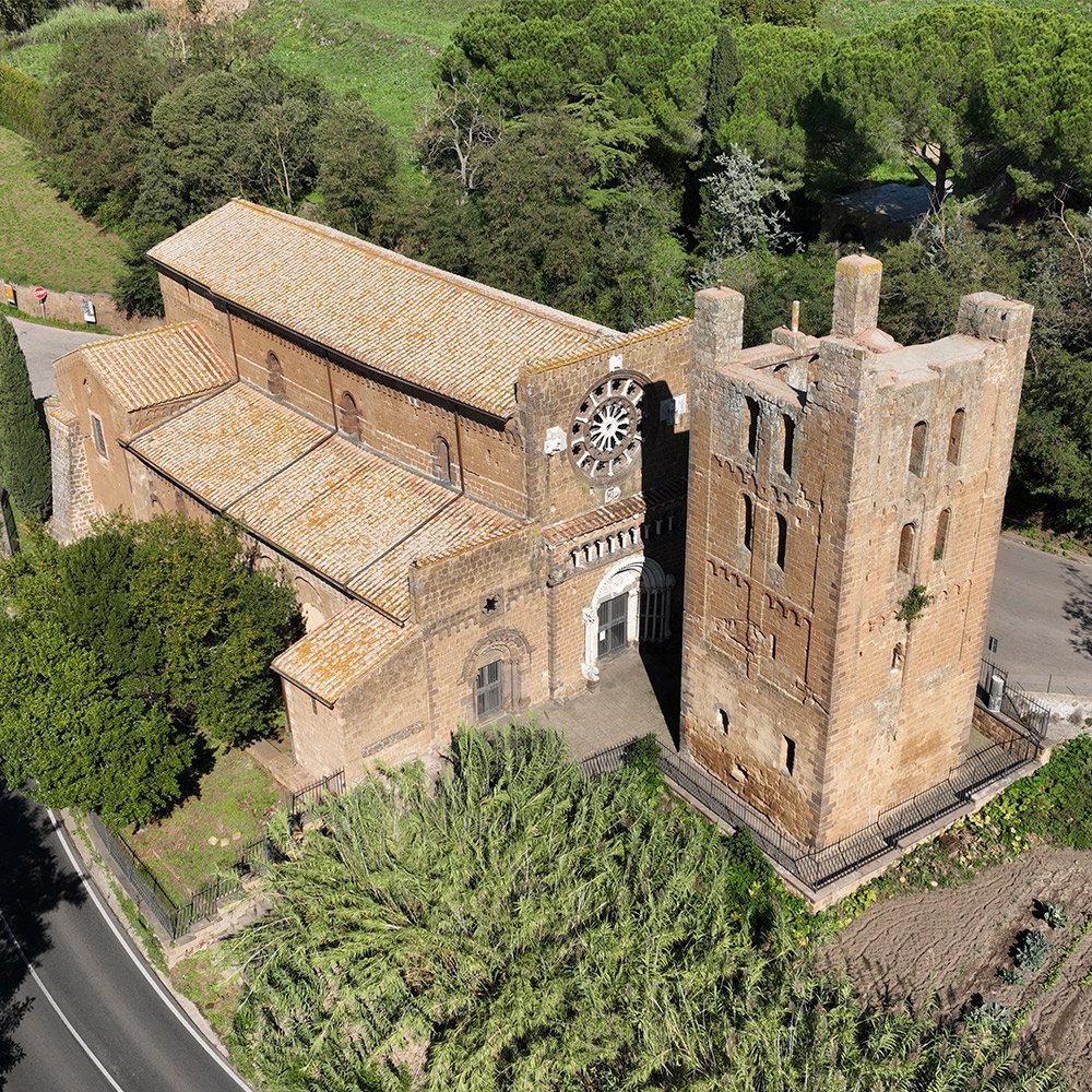 Un antico edificio in stile medievale con muro in pietra, una torre e un orologio, circondato da alberi verdi e coltivazioni.