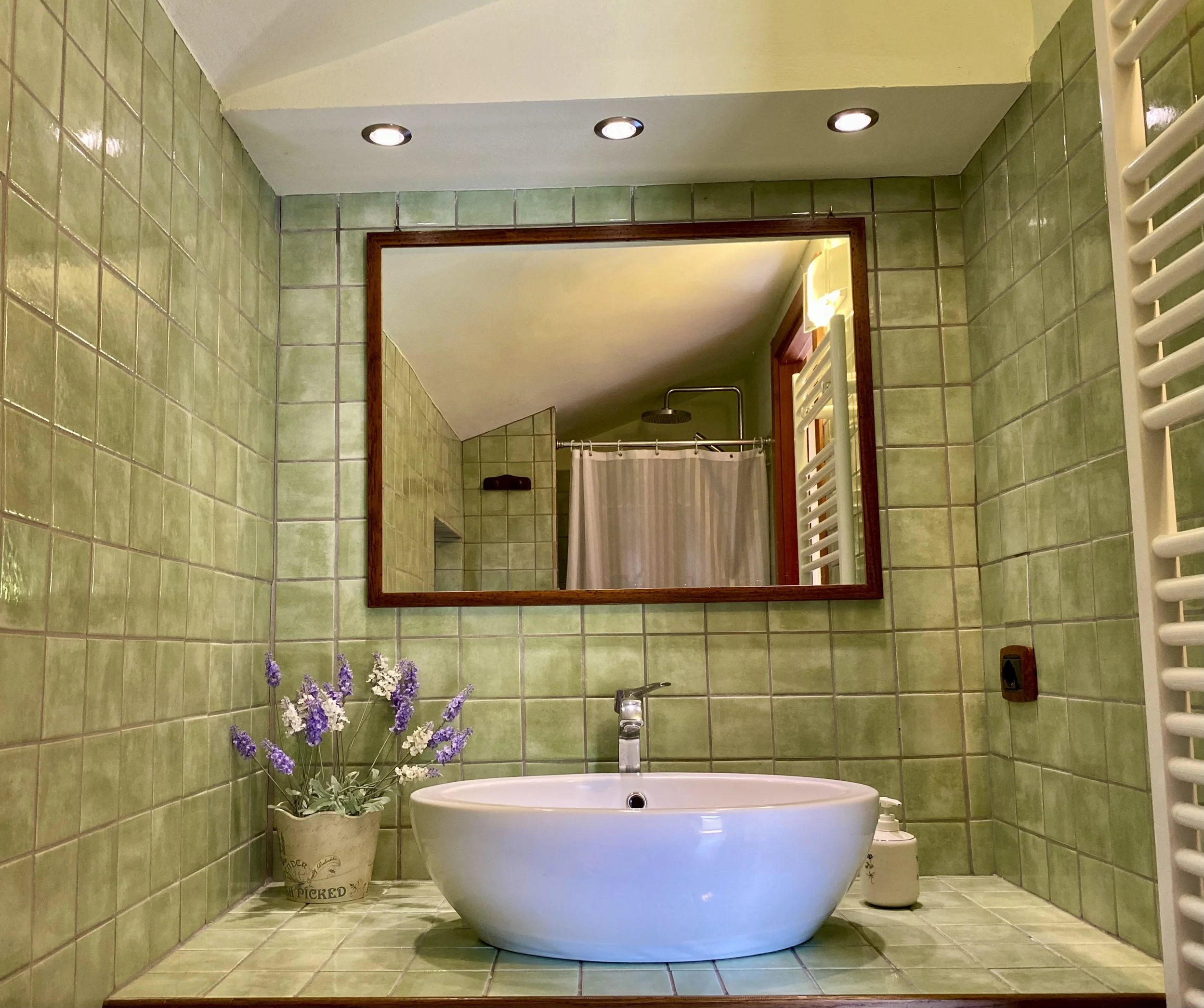 Bagno con pareti e pavimento rivestiti con piastrelle verdi, grande vasca da bagno bianca, vaso con fiori di lavanda e altri fiori, specchio con cornice di legno luminoso, soffitto con faretti, canna da riscaldamento e doccia con tendina.