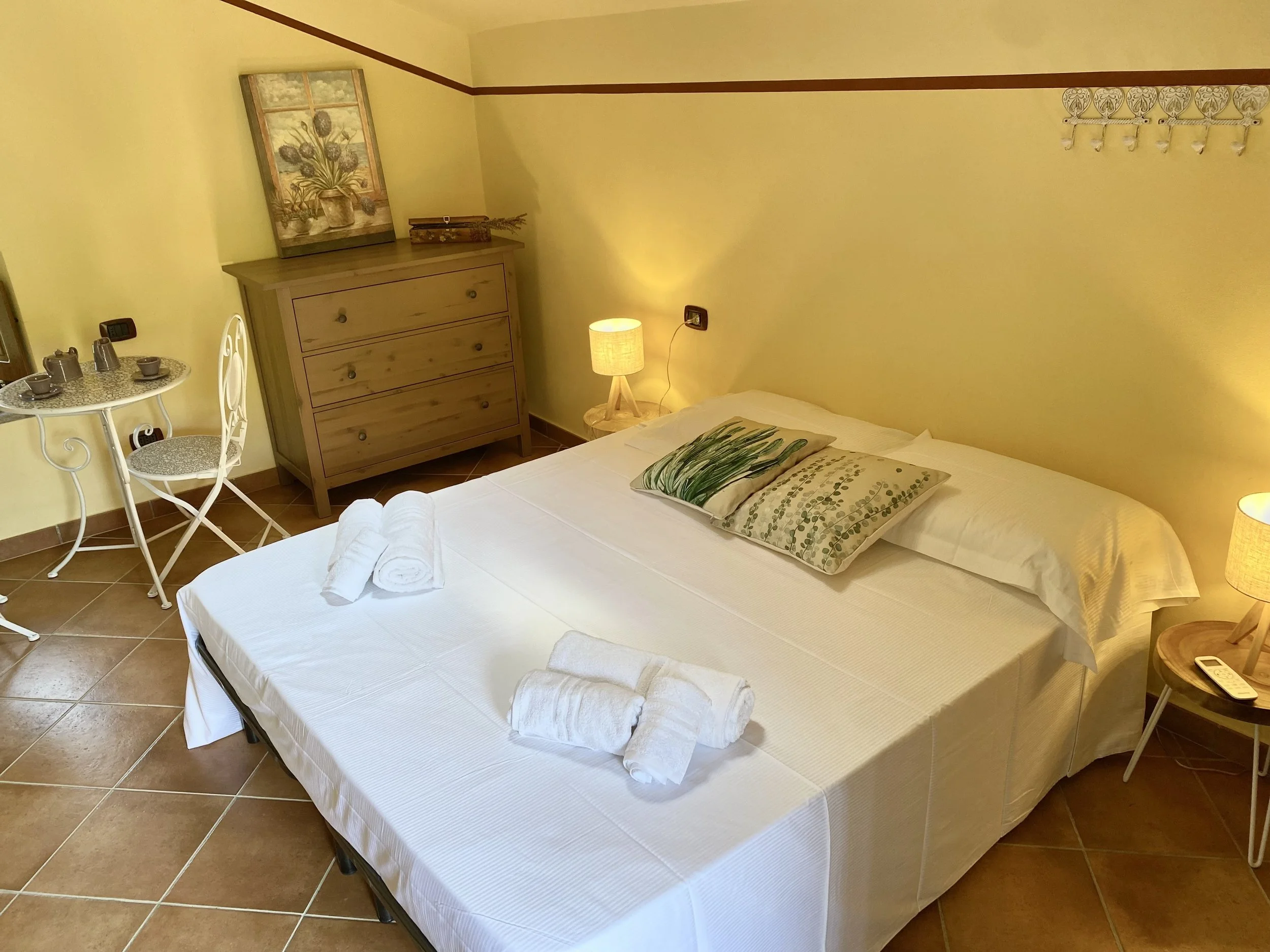 Camera da letto con letto matrimoniale, cuscini decorativi con foglie verdi, due asciugamani arrotolati sulle lenzuola, due lampade da tavolo accese, un comodino con telefono, una cassettiera in legno con sopra un quadro raffigurante fiori e piante, una piccola tavola con tazze, teiera e zuccheriera, una sedia bianca con sedile imbottito, pareti gialle con un quadro decorativo e appendiabiti con sei ganci.
