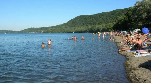 Spiaggia affollata con persone che nuotano e si rilassano sotto l'ombrellone, con un paesaggio montano sullo sfondo.