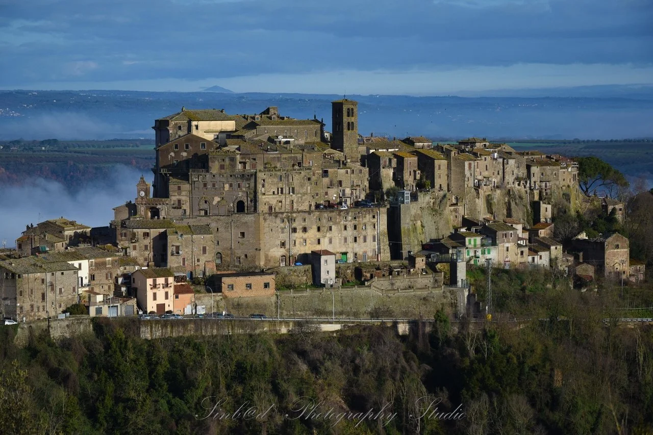 Antica città medievale su una collina, con edifici in pietra e torre campanaria, circondata da colline verdi e un cielo nuvoloso.