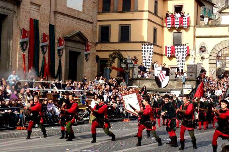Una parata con persone in costumi tradizionali in strada, con un pubblico che osserva e decorazioni storiche sullo sfondo.