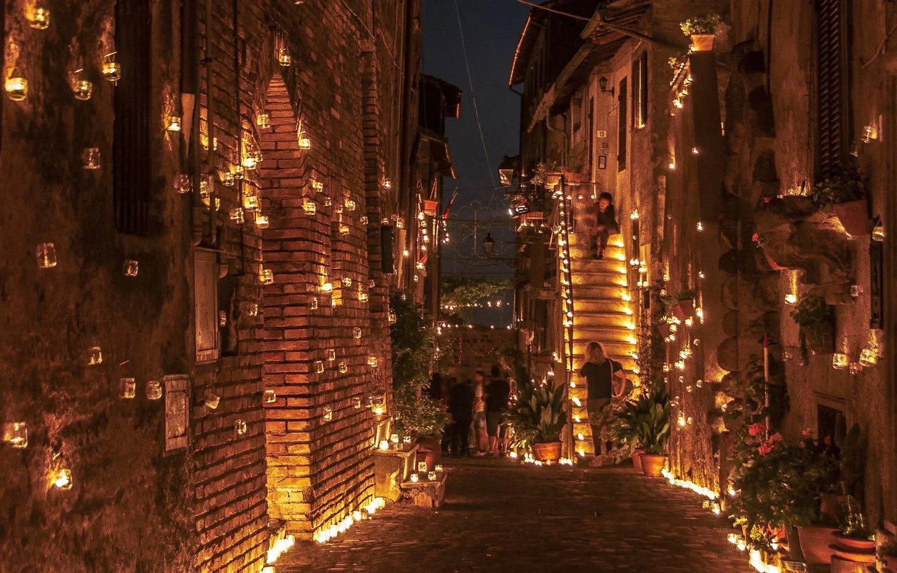 Una strada di notte illuminata da numerosi lumini appesi alle pareti di edifici e lungo i gradini di una scala, con alcune persone che camminano e piante in vaso.