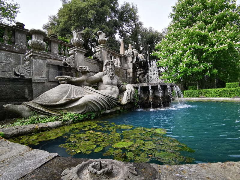 Statua di un uomo seduto, con un'espressione seria, accanto a una fontana con acqua che scorre, circondata da alberi e vegetazione, in un giardino.