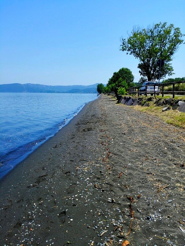 Spiaggia con sabbia scura, bottiglie di vetro, ciottoli e detriti sulla riva di un lago o mare. Sullo sfondo, alberi e una strada con un'autoferma.