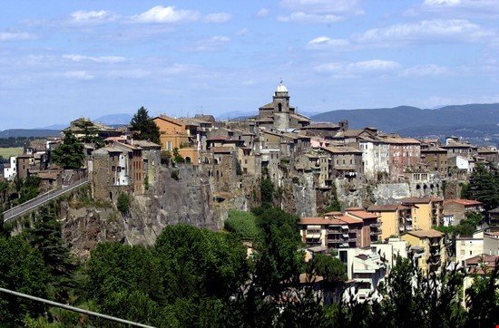Paesaggio di un paese collinare con edifici storici e un castello in cima, circondato da natura e montagne lontane.