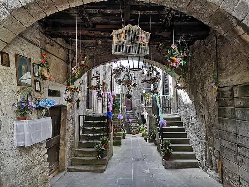 Ingresso di un vicolo in pietra con scale decorate con fiori e piante, archi in pietra e decorazioni sospese.
