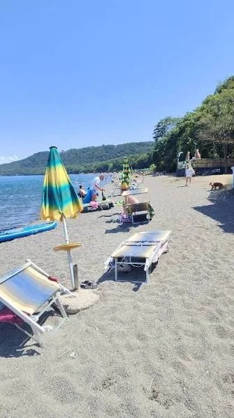 Spiaggia con ombrelloni, lettini e persone che si rilassano vicino al lago o al mare, con albero e montagne sullo sfondo.