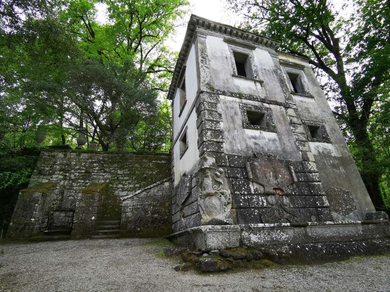 Edificio abbandonato in stile antico, con pareti bianche e finestre senza vetri, circondato da alberi verdi, con statue di pietra e scale in pietra ai lati.