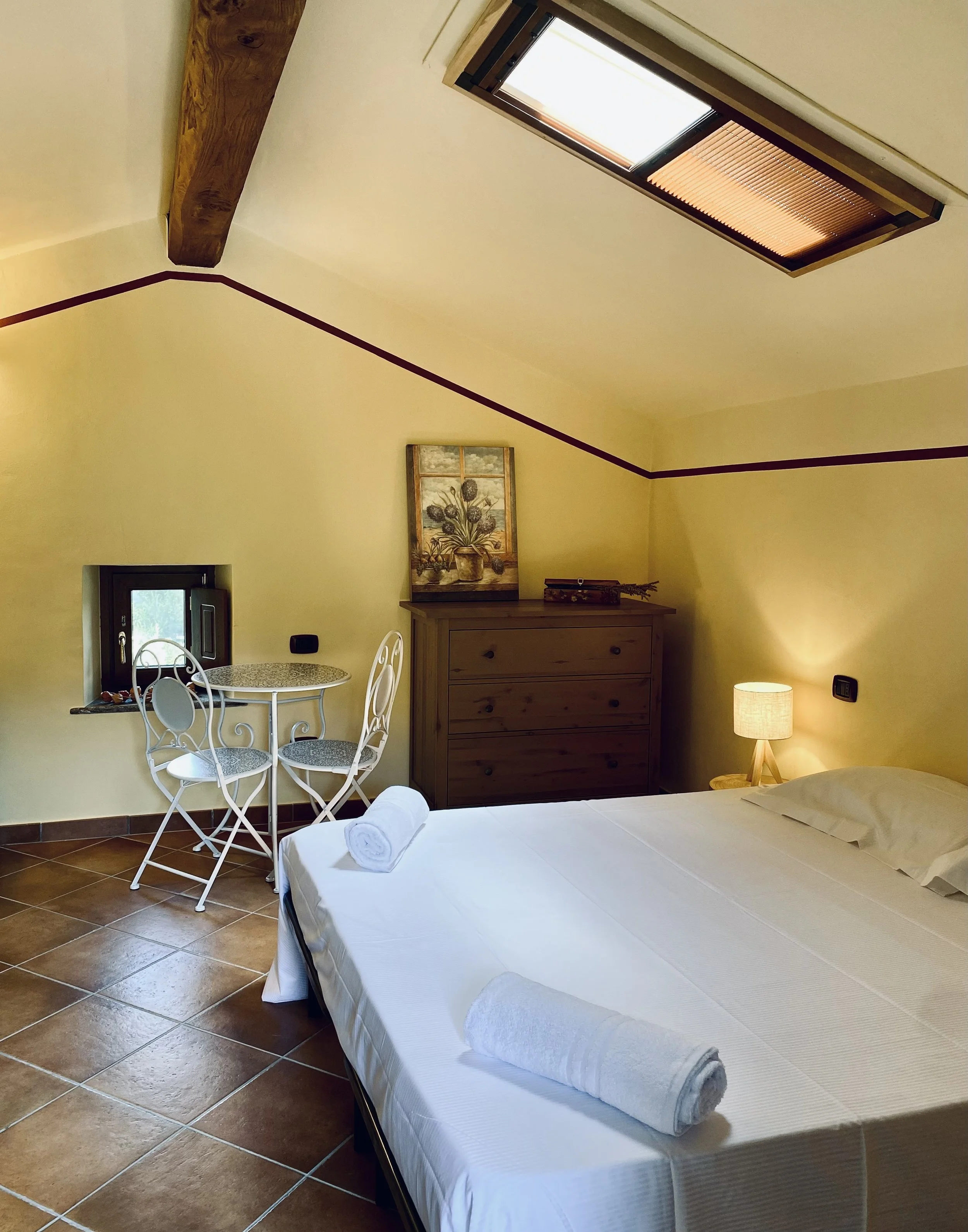 Camera da letto con letto matrimoniale con lenzuola bianche e asciugamani arrotolati, lampada da tavolo accesa, finestra con tende veneziane e soffitto con finestra da cui entra luce naturale, pareti gialle decorate con quadro di fiori, tavolo rotondo con sedie di ferro battuto, cassettiera di legno, e pareti con linea decorativa viola.