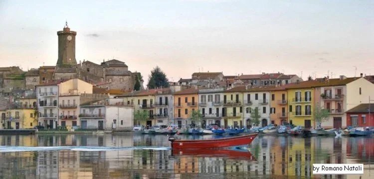 Veduta di un villaggio di case colorate lungo un fiume, con barche attraccate, e un castello con torre sulla collina sullo sfondo.
