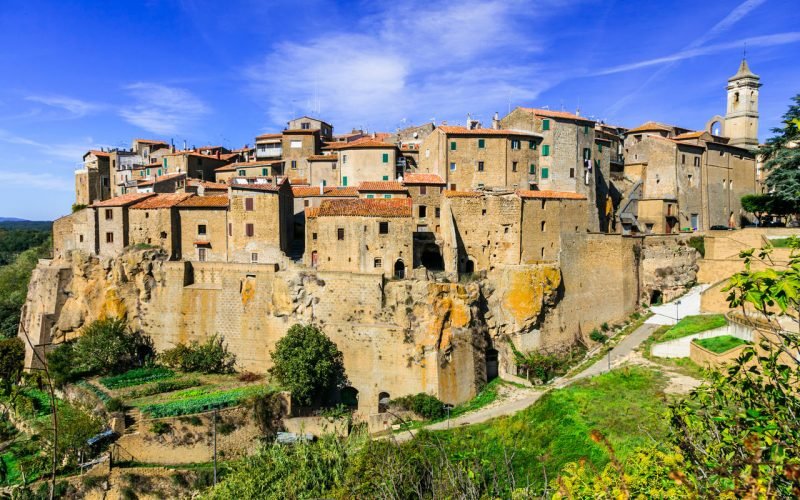 Paesaggio con un insediamento storicamente costruito su un promontorio roccioso, composto da case con tetti rossi e una chiesa con un campanile bianco in alto a destra.
