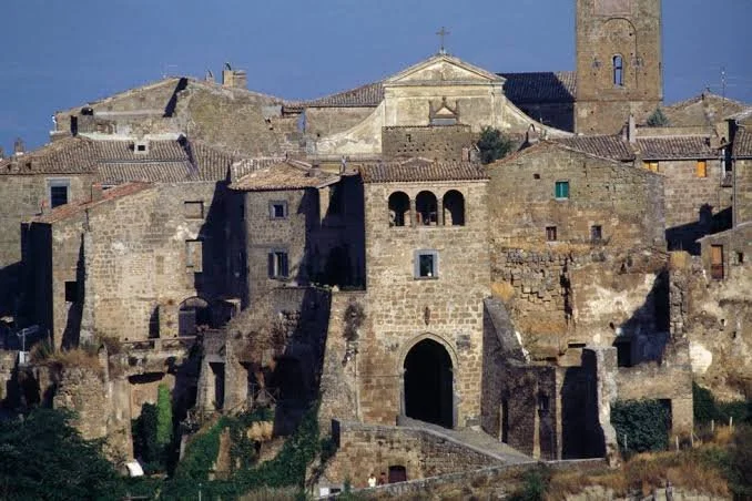 Vista di un antico complesso di edifici in pietra con una chiesa in cima, in un paesaggio collinare, con torri e case storiche.