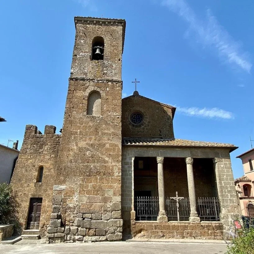 Chiesa antica in pietra con campanile e portico con colonne, su sfondo di cielo blu.