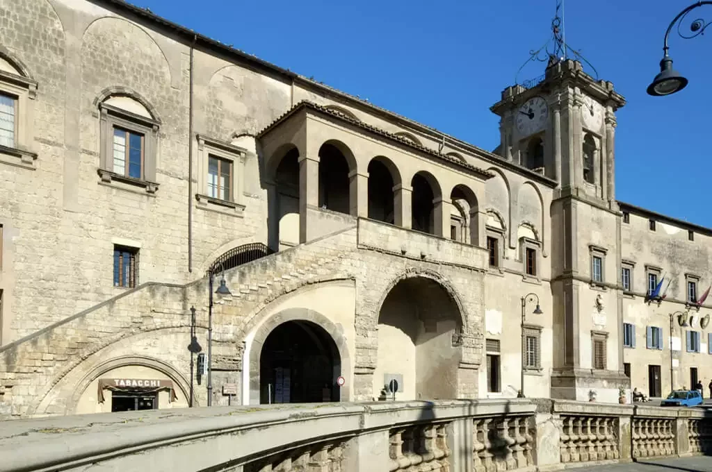 Edificio storico con arcate e un campanile davanti a un cielo azzurro, con lampioni e alcune bandiere appese lungo la facciata.