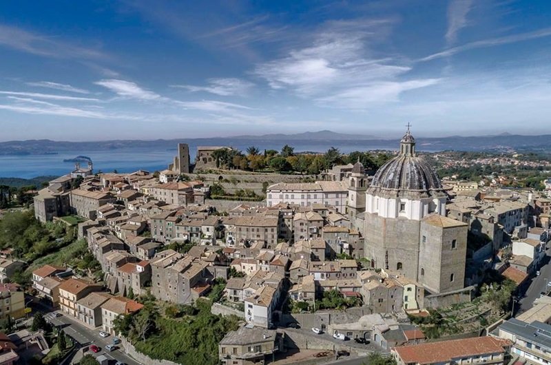 Veduta aerea di un antico borgo medievale con case in pietra, una grande chiesa con cupola e un castello, vicino a un lago e montagne sullo sfondo.