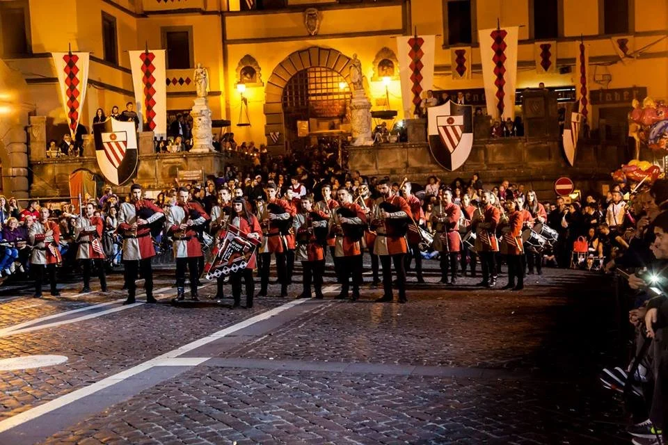 Una band musicale in uniforme rossa e nera suona in una piazza storica, con decorazioni e bandiere medievali appese agli edifici circostanti, in una scena notturna.