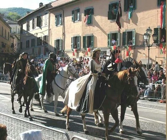 Persone in abiti storici a cavallo partecipano a una sfilata in una strada di un paese con edifici colorati e spettatori al bordo della strada.