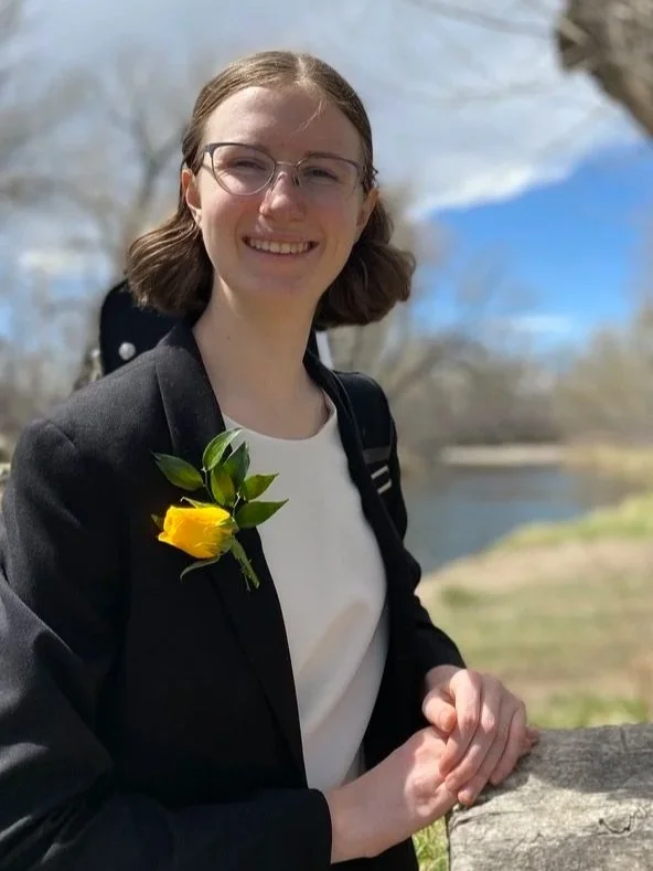 A young woman with glasses and shoulder-length hair smiling outdoors. She is wearing a black blazer with a yellow flower pin on it, a white shirt, and has a backpack. The background shows a river, trees, blue sky, and clouds.