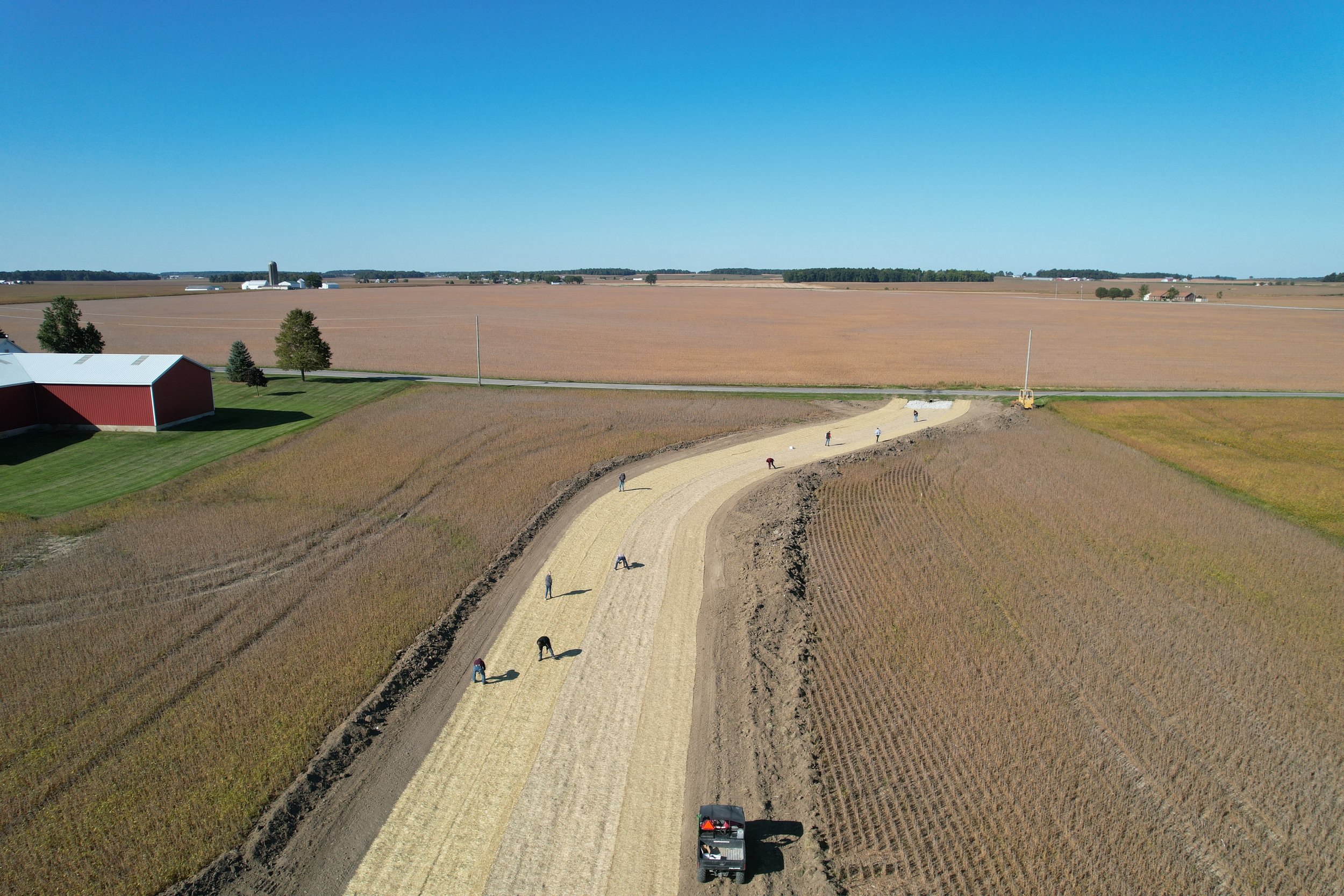 Aerial view of a farm with a red barn, fields, and a dirt road under construction with workers and construction vehicles.