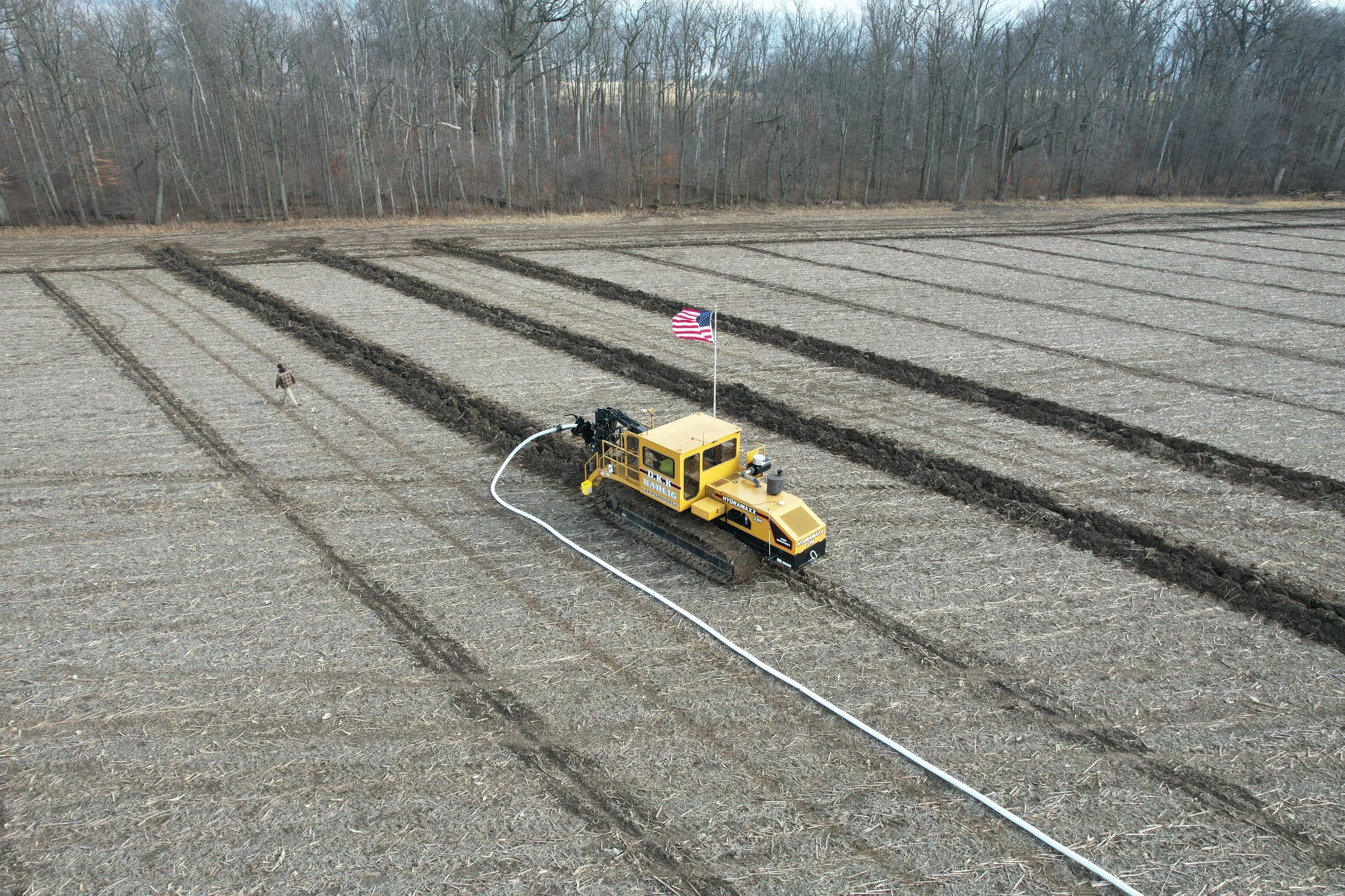 A yellow farm irrigation machine watering a large field with furrows, there is a person walking nearby, and a small American flag attached to the machine. In the background, there is a line of leafless trees.