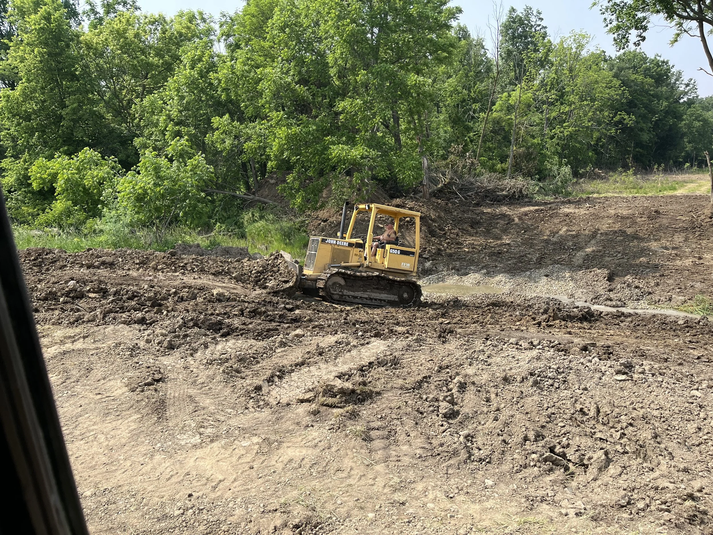 A yellow John Deere bulldozer moving dirt on a construction site with lush green trees in the background.
