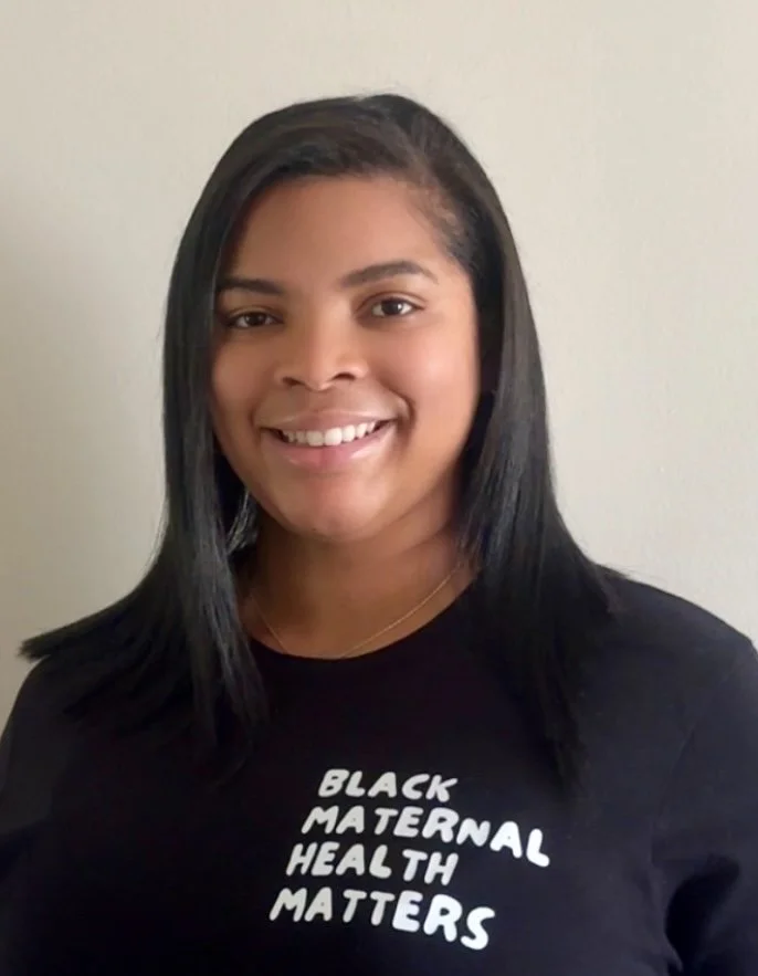 A young woman with straight black hair smiling at the camera, wearing a black t-shirt that says "Black Maternal Health Matters."