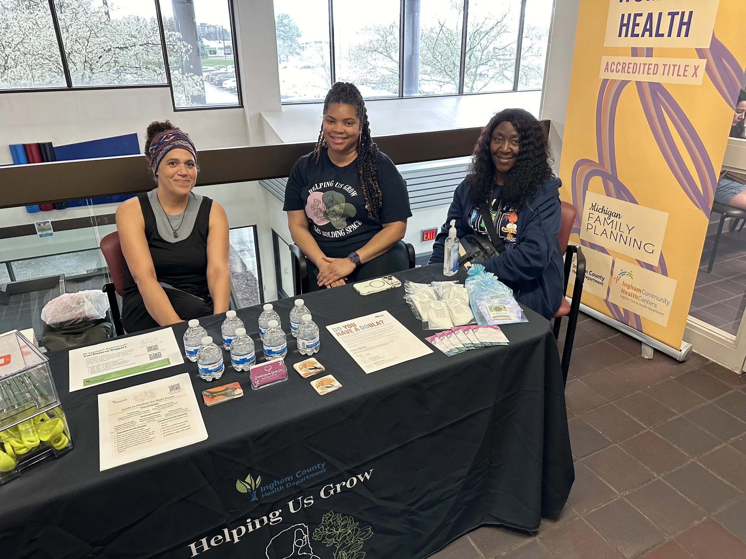 Three women sitting at an information booth table with water bottles and informational pamphlets, inside a building with large windows and snowy trees outside, purple and yellow banners promoting health and family planning in the background.
