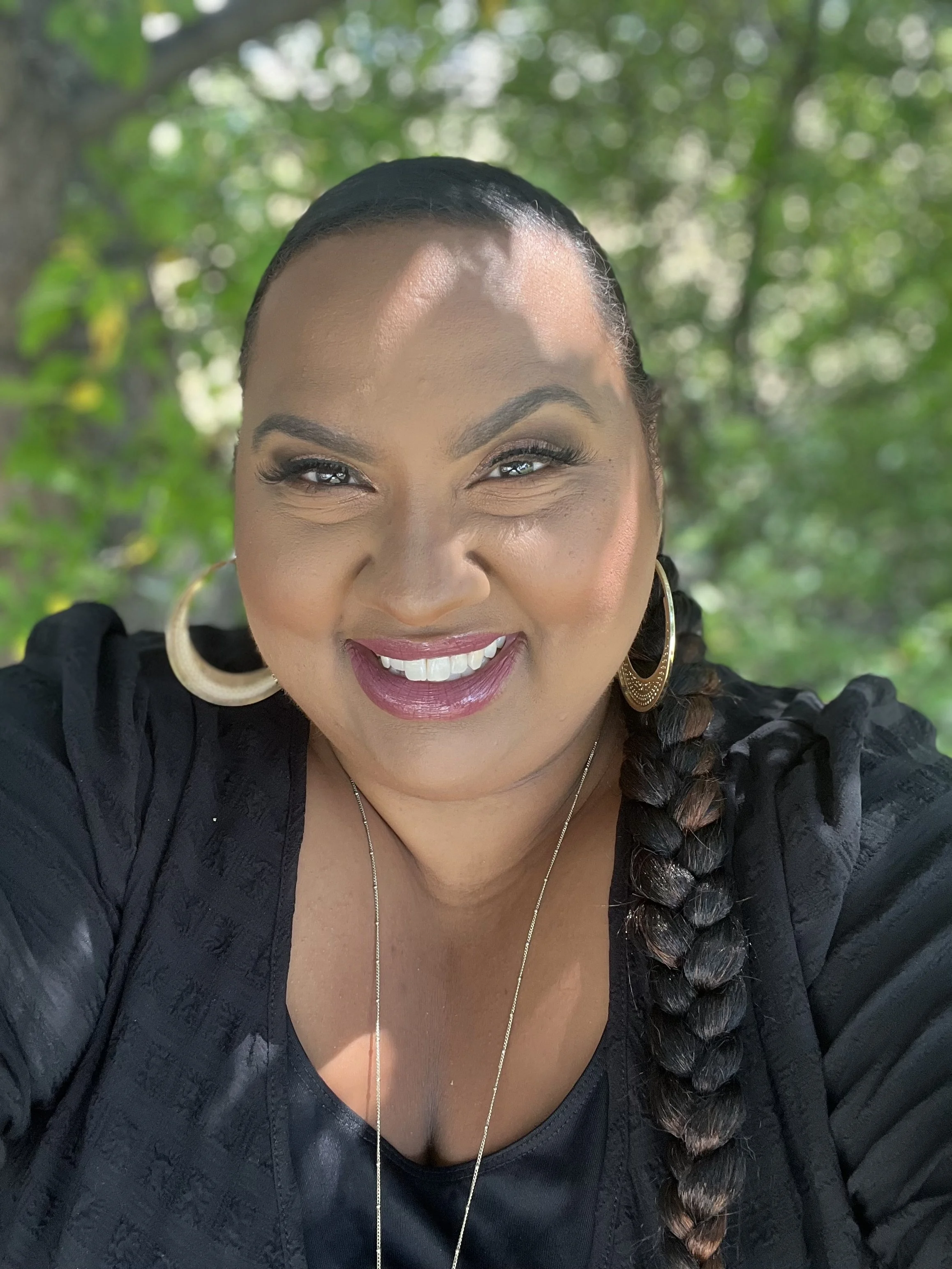 Close-up of a smiling woman with braided hair, wearing large hoop earrings and a black textured top, outdoors with a blurred green leafy background.