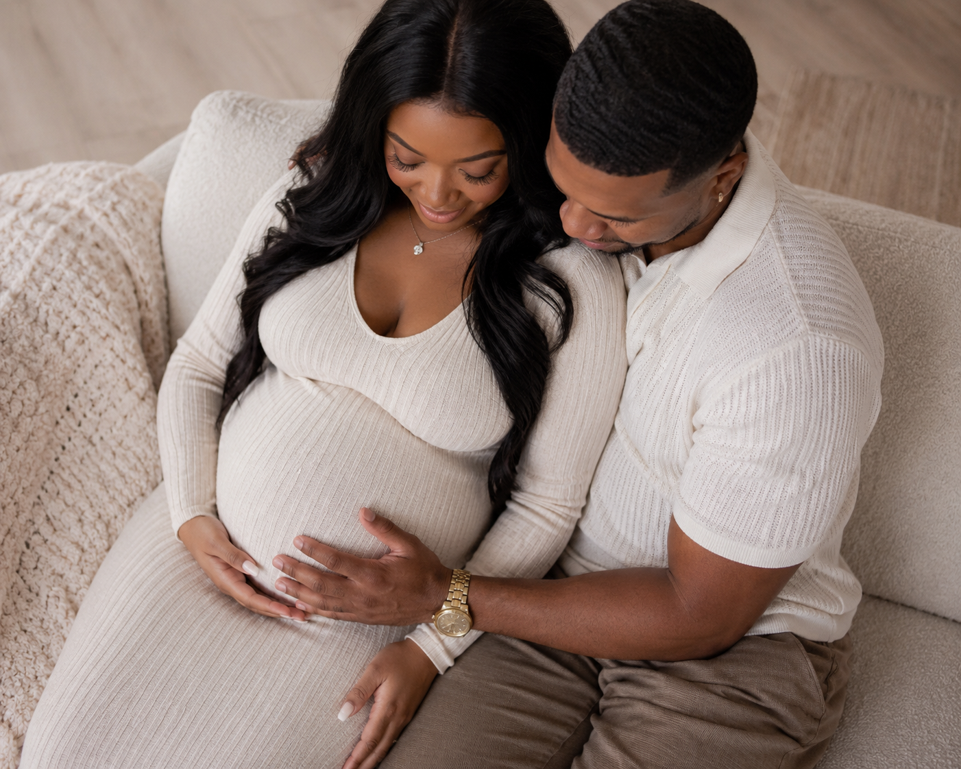 A pregnant woman sits on a beige couch, gently holding her belly, next to a man who is touching her belly and looking down affectionately, both wearing matching beige outfits.