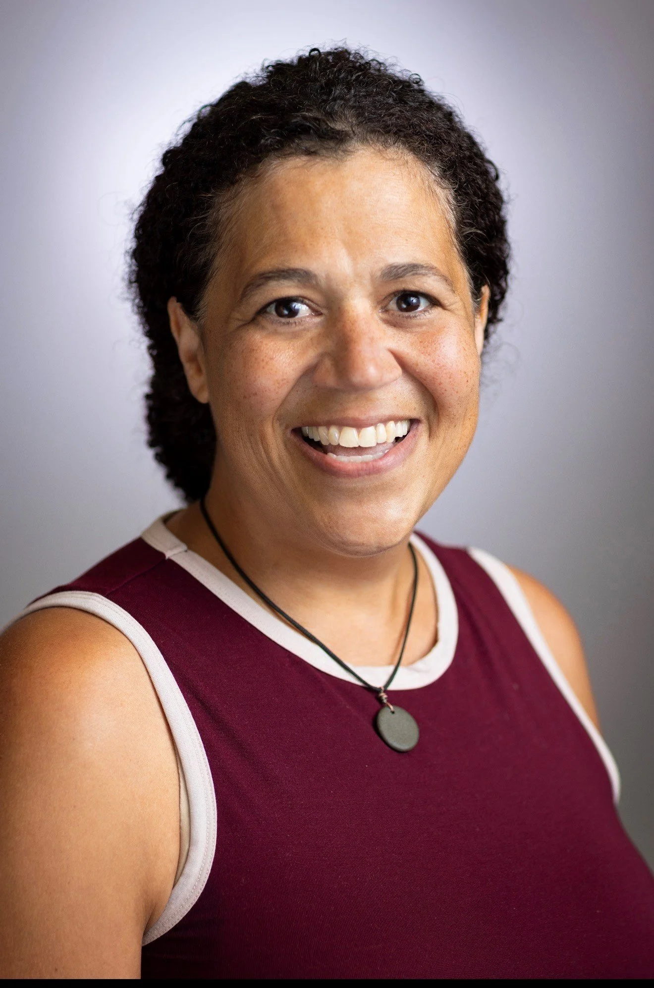 A woman with curly dark hair, smiling with teeth showing, wearing a sleeveless maroon top with light-colored trim, a round pendant necklace, and a black choker, standing against a plain light background.