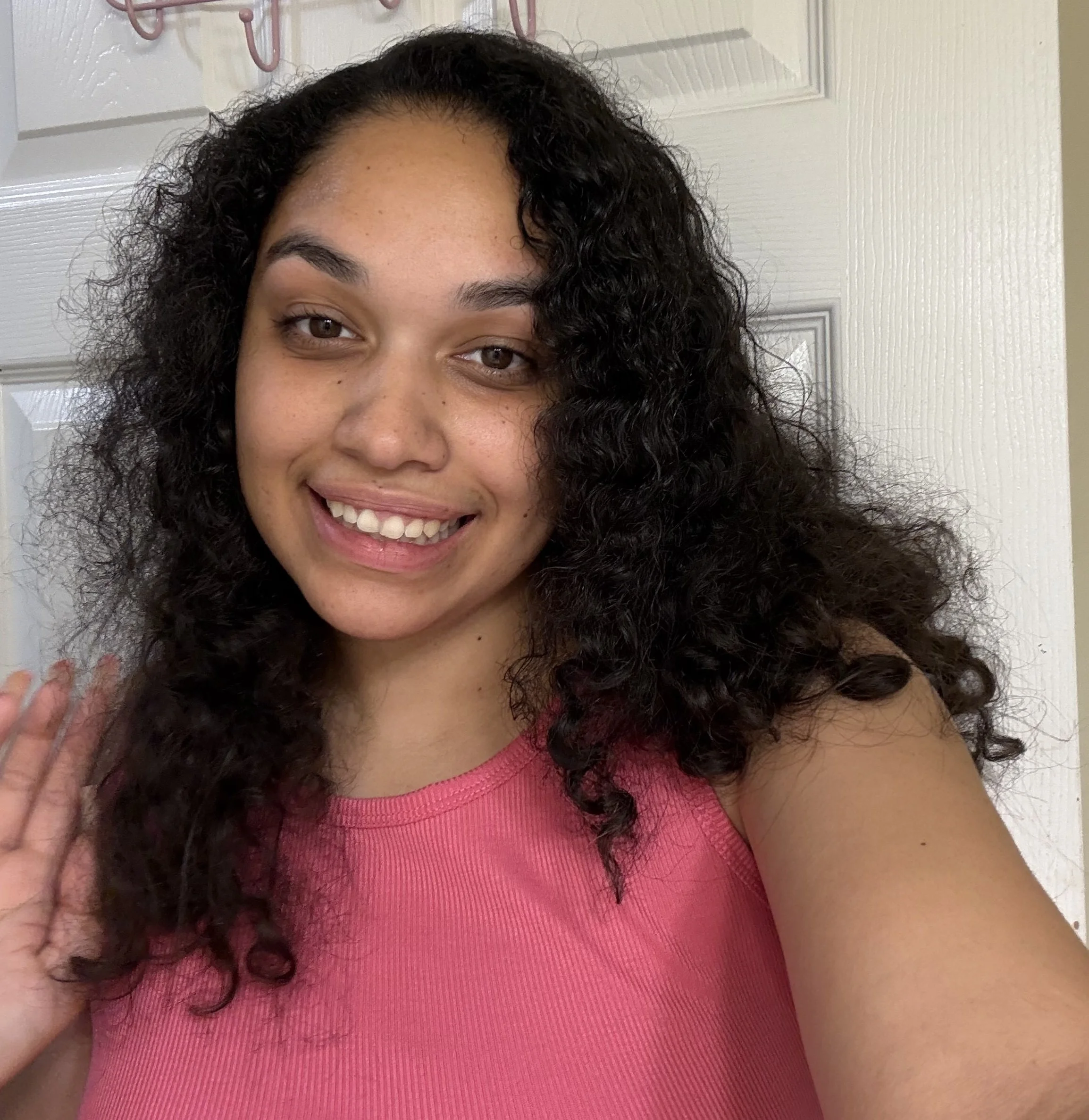 Close-up image of a woman with curly dark hair, wearing a pink sleeveless top, smiling at the camera, with a light-colored door in the background.