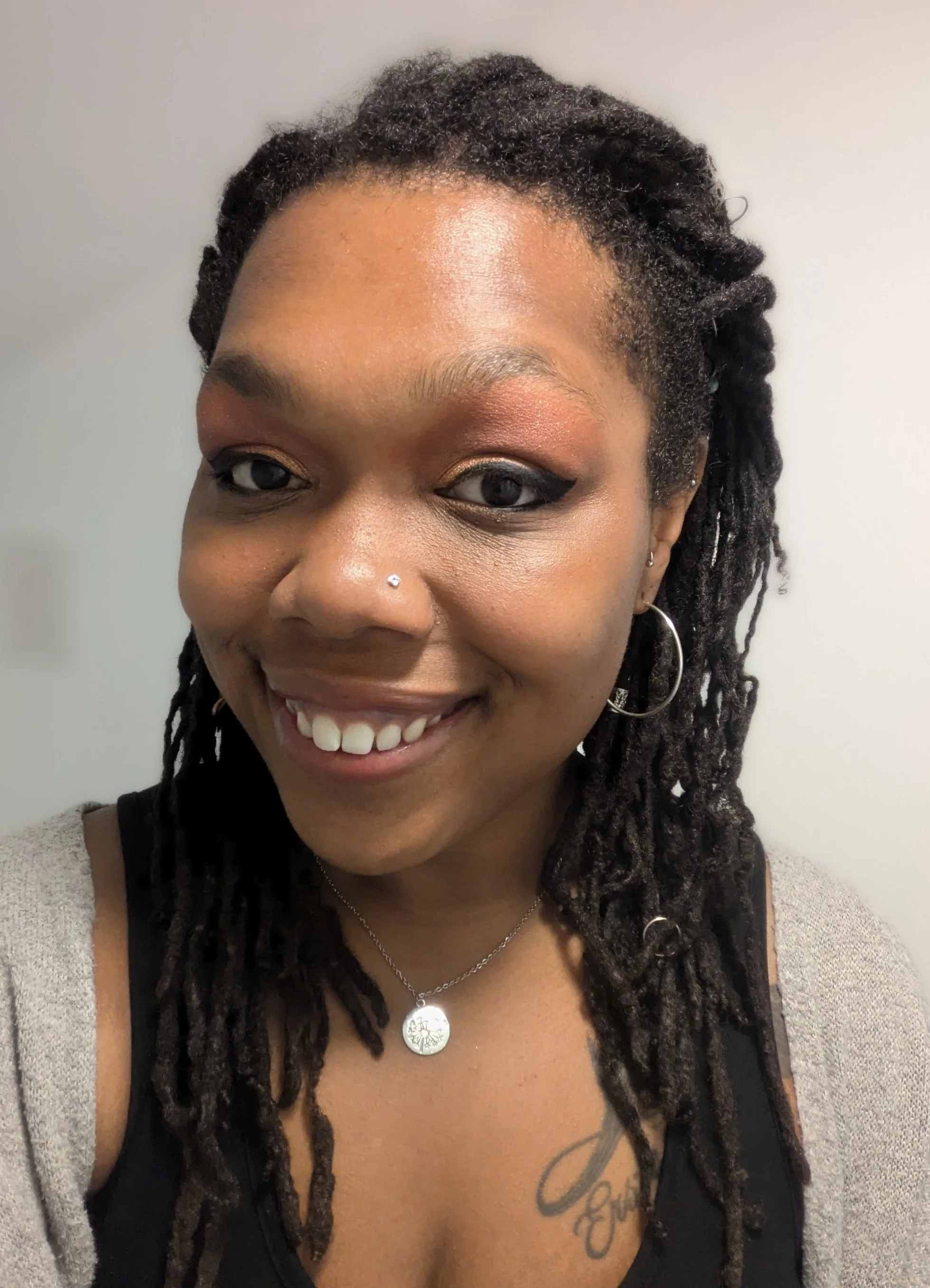 A smiling woman with dark dreadlocks, wearing a gray cardigan, black top, silver necklace, and hoop earrings.