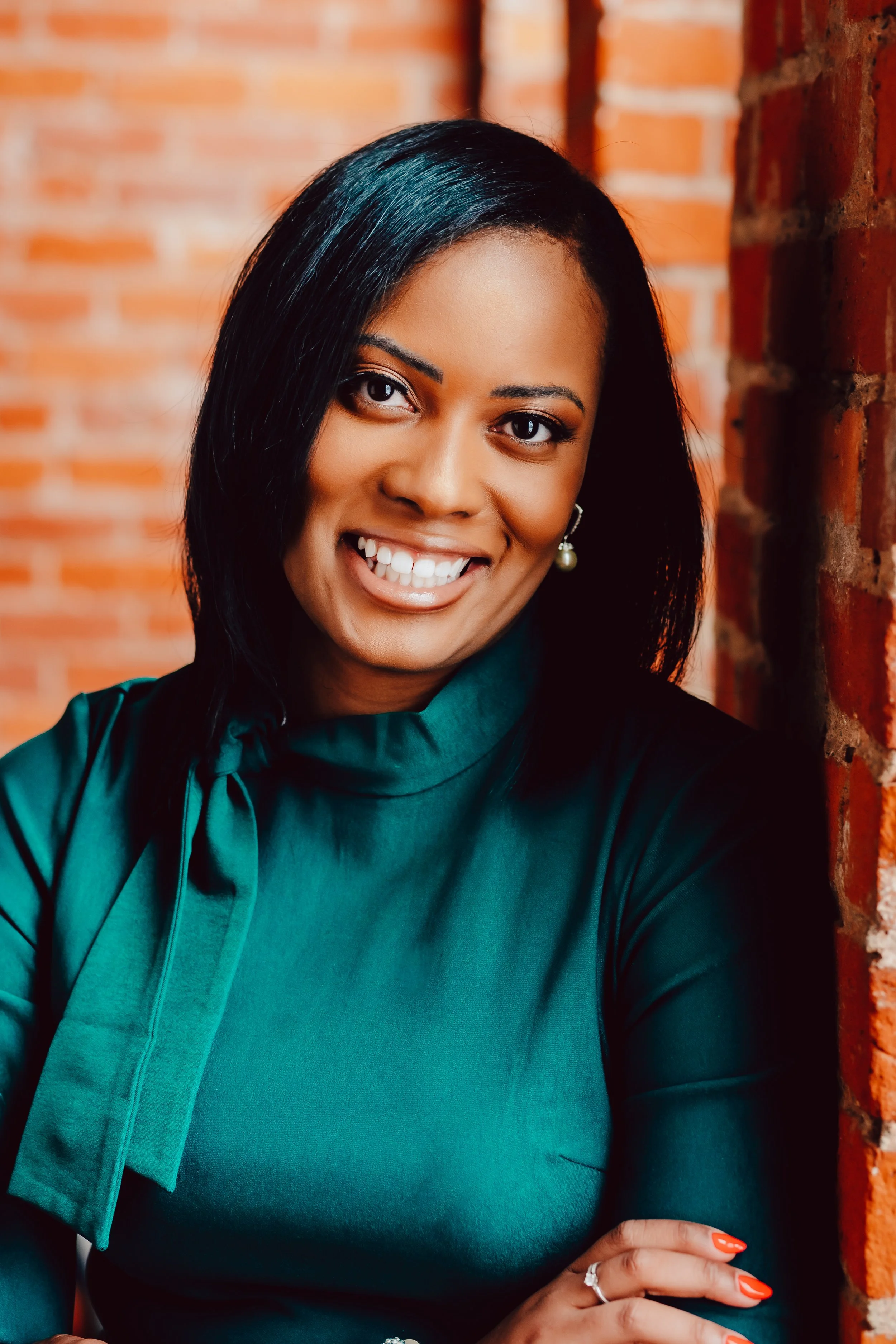 Close-up of a smiling woman with dark hair, wearing a teal blouse, standing against a brick wall.