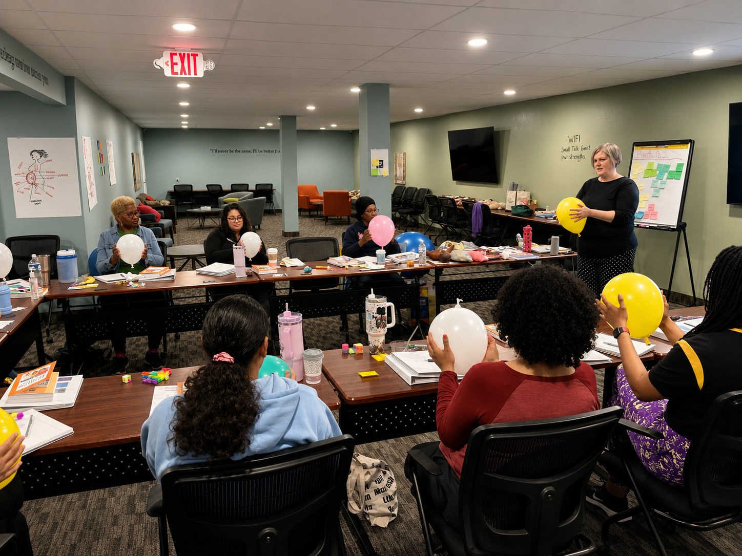 A woman leading a doula workshop with balloons at a conference room, seated participants holding balloons, notebooks, and pens, with a whiteboard and TV in the background.