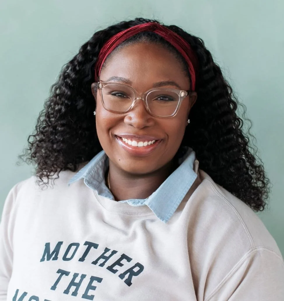 A smiling woman with glasses and curly hair, wearing a headband and a sweatshirt that says 'Mother the', in front of a light green background.