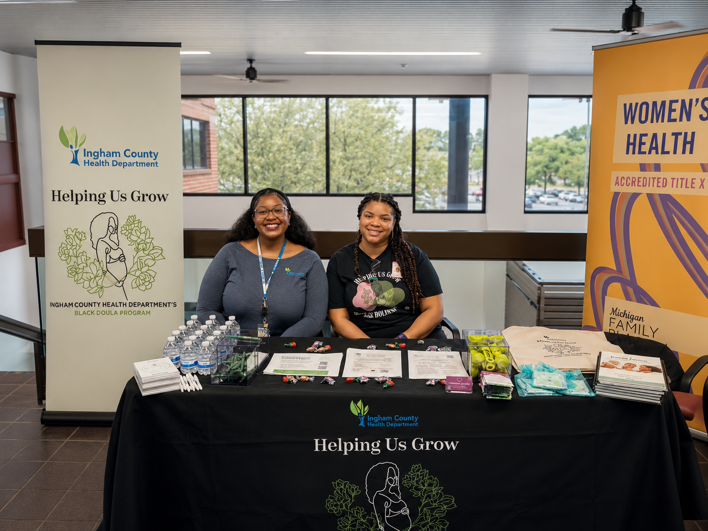 Two women sitting behind a table at a doula event, smiling. Behind them are banners for Ingham County Health Department and Women's Health.