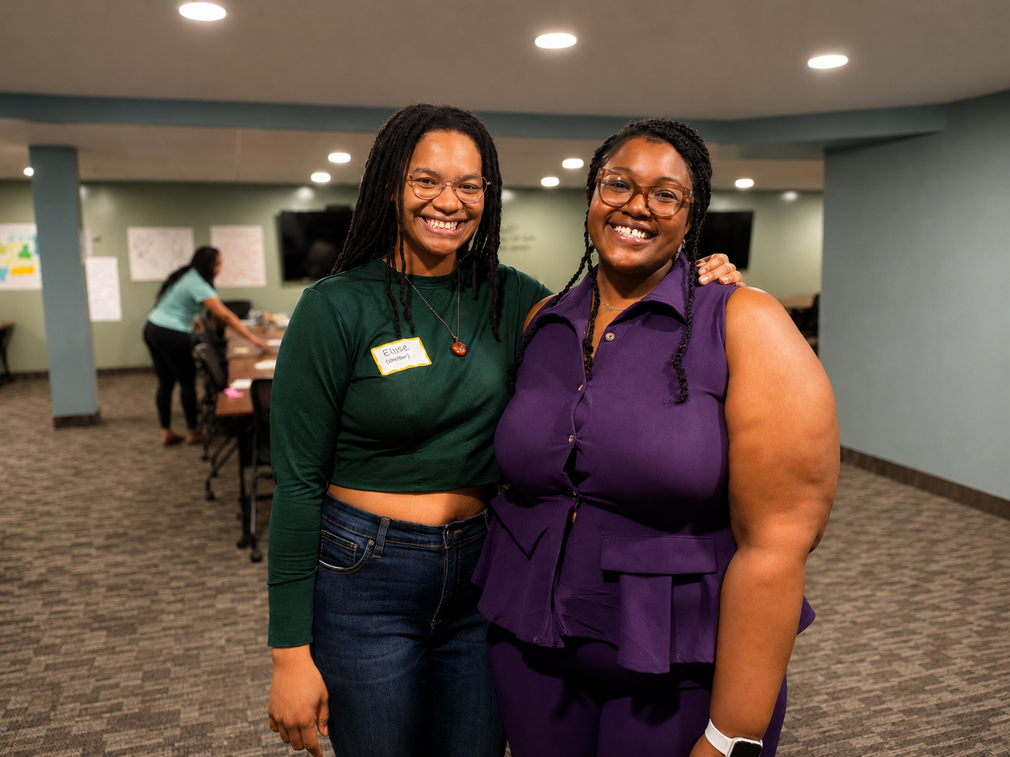 Two women smiling and posing in an indoor setting, one is wearing a green top and glasses, the other is wearing a purple sleeveless dress and glasses, with a woman in the background working at a table.